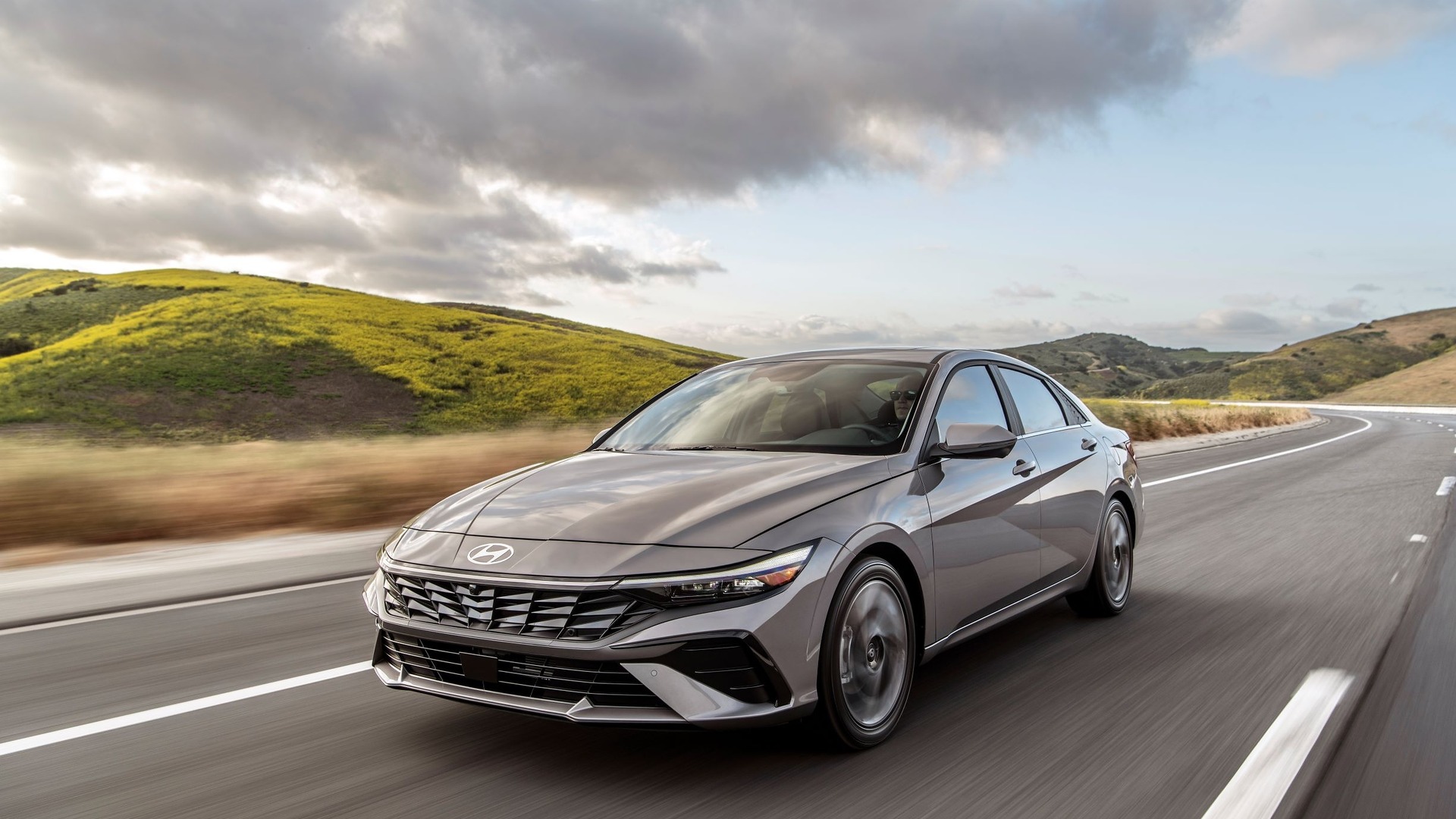 Elantra 2024 restylée argent, vue trois-quarts avant en conduite sur route de collines | Hyundai Elantra 2024 restylée argent, vue trois-quarts avant en conduite sur route de collines