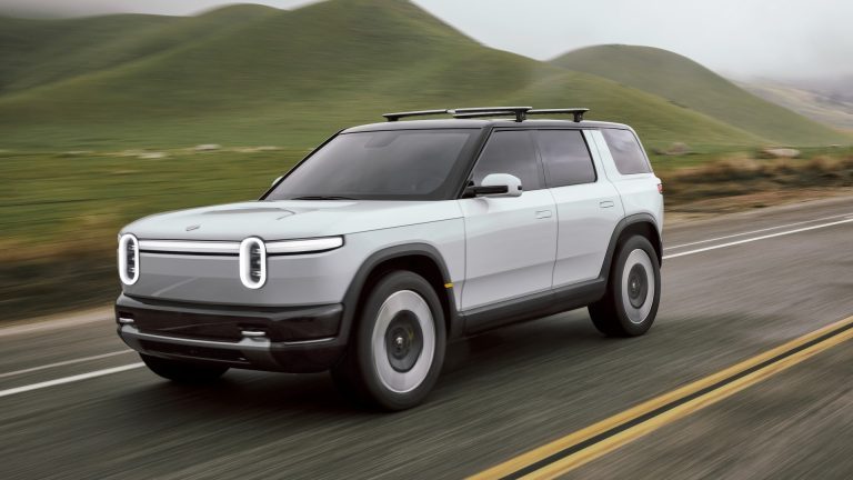 Rivian R2 de présentation 2024, gris pâle, roulant sur une route de campagne bordée de collines, vu de trois-quarts avant