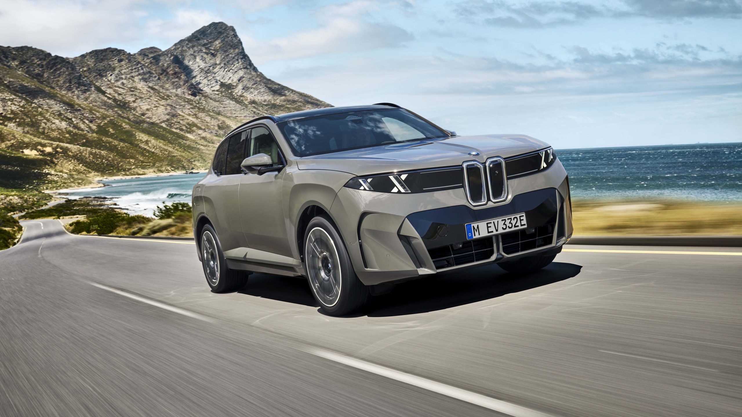 2027 BMW iX5 60 xDrive electric SUV in silver driving along a scenic coastal road, with rugged mountains, turquoise ocean and blue skies in the background.