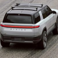 Rivian R2 de présentation 2024 vu de trois-quarts arrière en plongée, avec son bandeau lumineux arrière et ses barres de toit | Rivian Rivian R2 de présentation 2024, gris pâle à toit noir, roulant sur une route, vu de haut et de trois-quarts arrière