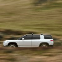 Rivian R2 de présentation 2024, blanc, de profil en mouvement sur une route de campagne | Rivian Rivian R2 de présentation 2024, blanc à toit noir, vu de profil complet en mouvement devant un champ