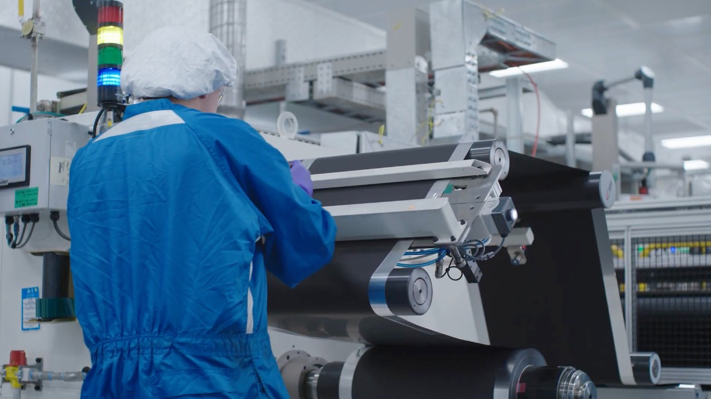 Technicien en combinaison bleue de salle blanche et charlotte, travaillant sur une machine à rouleaux de matériau noir dans un laboratoire de batteries