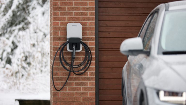 Borne de recharge résidentielle de niveau 2 fixée à un mur de briques près d’un garage, câble enroulé autour du support, avec une cour enneigée et un VUS électrique stationné